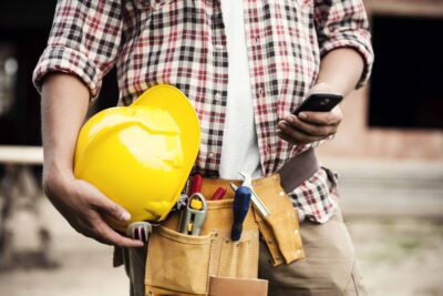 Man wearing a tool belt holding hard holding a cell phone.