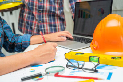 Desk with laptop, hard hat, glasses, magnifying glass and two people at it