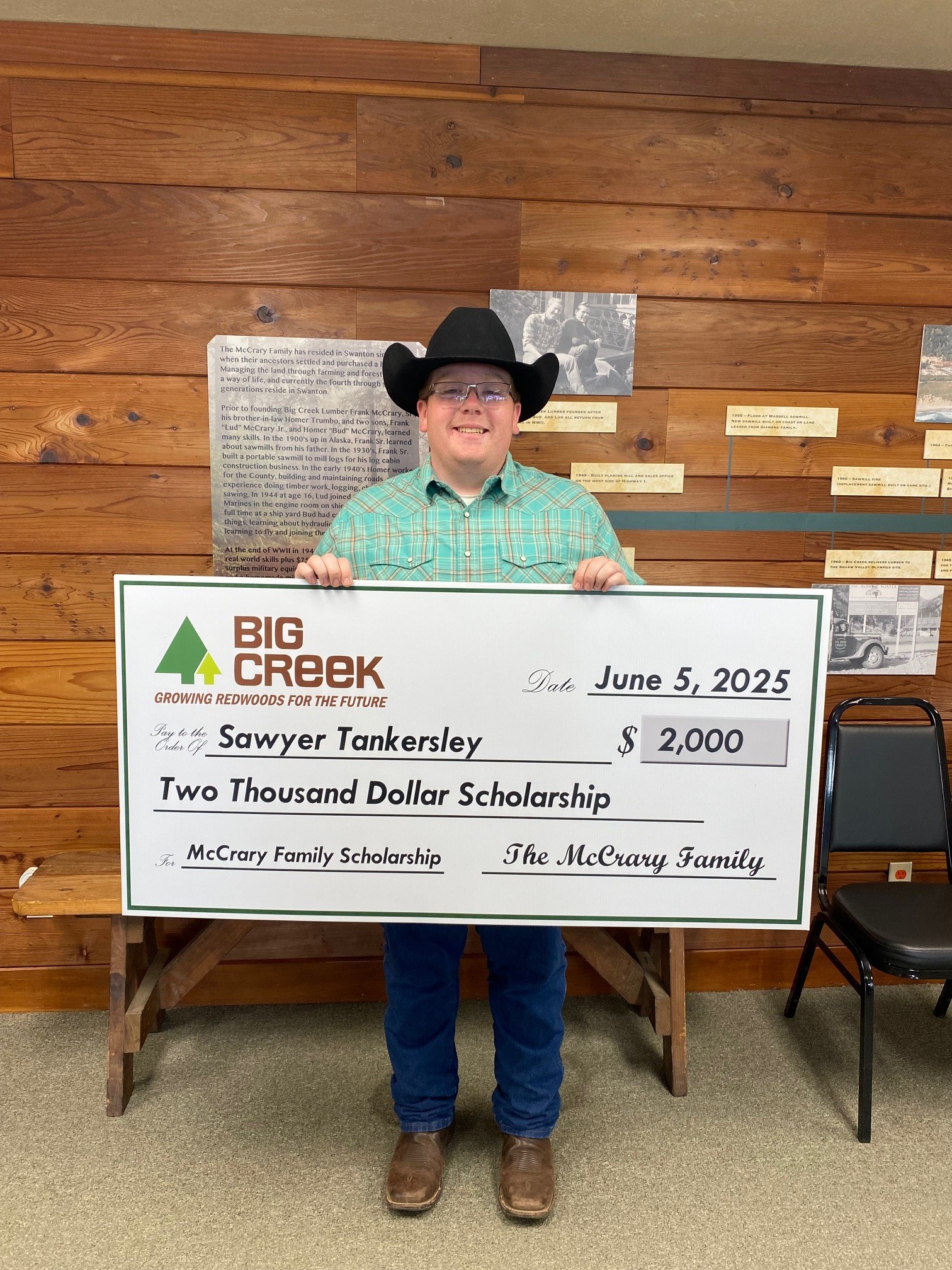 Young man with a cowboy hat standing with scholarship check in front of a wooden wall